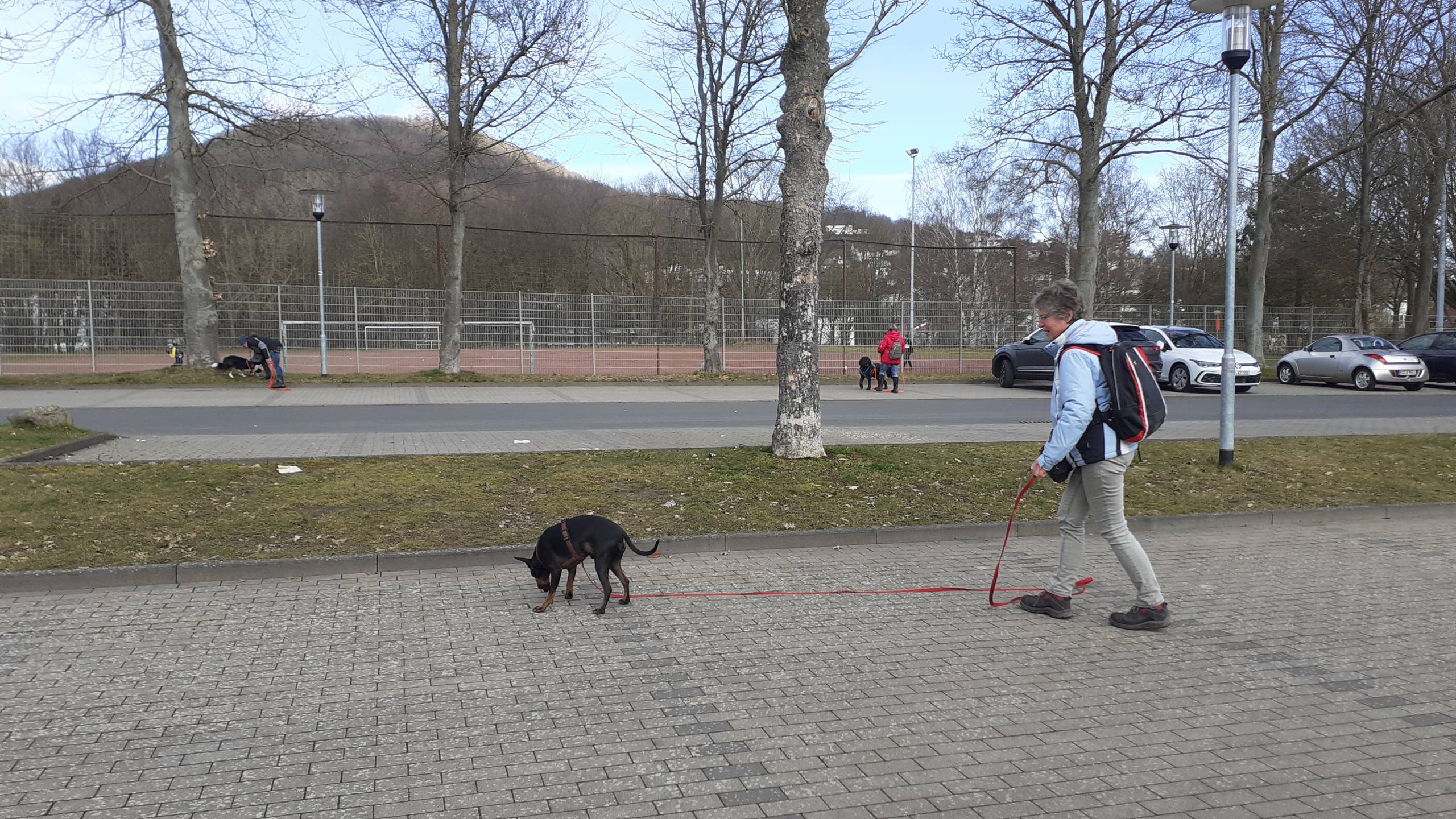 Frau geht mit ihrem Hund an der Leine an einem Sportplatz entlang.